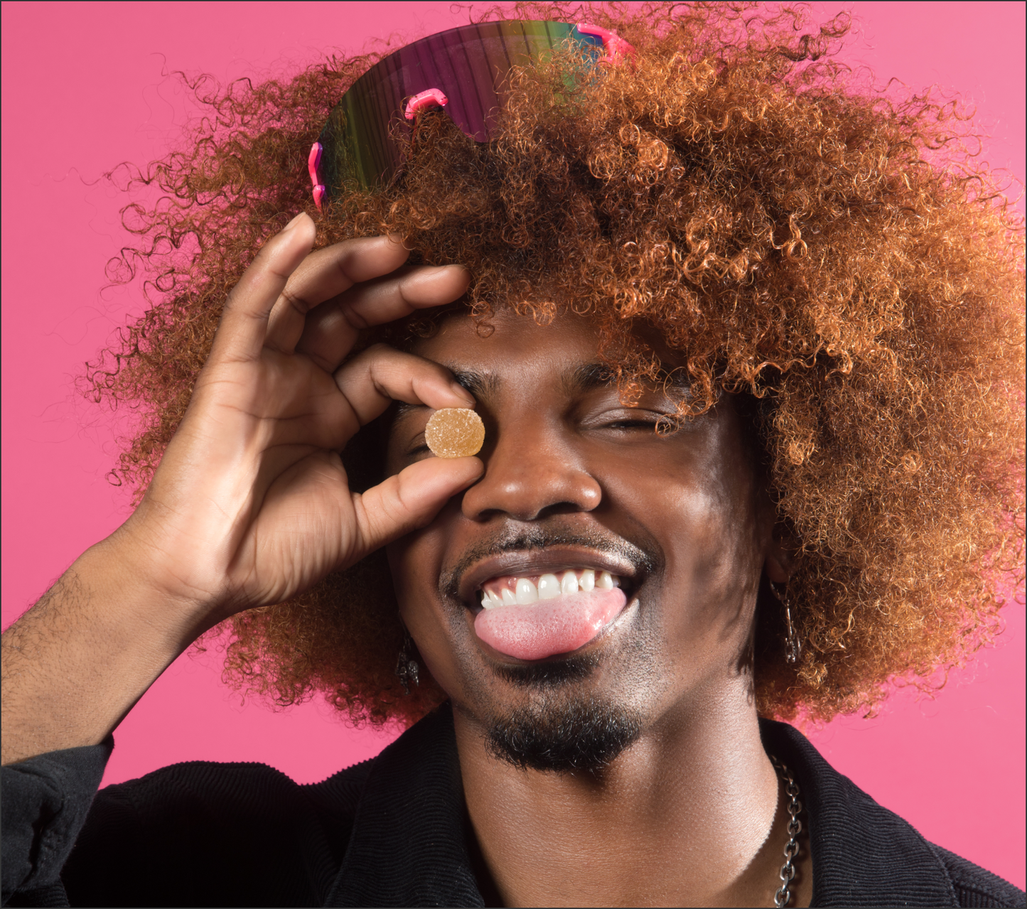 Smiling person with curly hair holding a round gummy over one eye against a pink background
