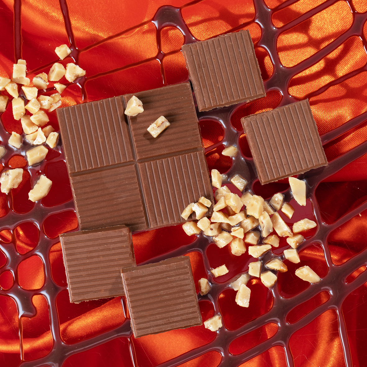 Milk chocolate squares and chopped nuts drizzled with chocolate sauce on red satin fabric.
