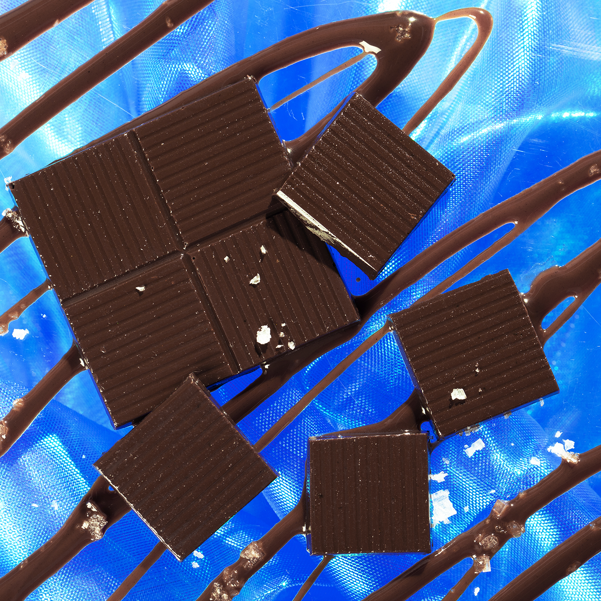 Dark chocolate squares with ridged tops, sea salt flakes and chocolate drizzle on a shiny blue surface