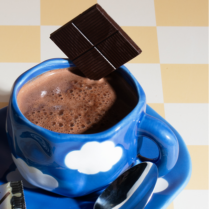 Blue cloud-pattern mug of frothy hot chocolate with a spoon and a square of dark chocolate held above the mug