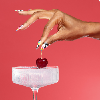 Hand with rainbow nail art holding a cherry by its stem above a glass compote against a red background.
