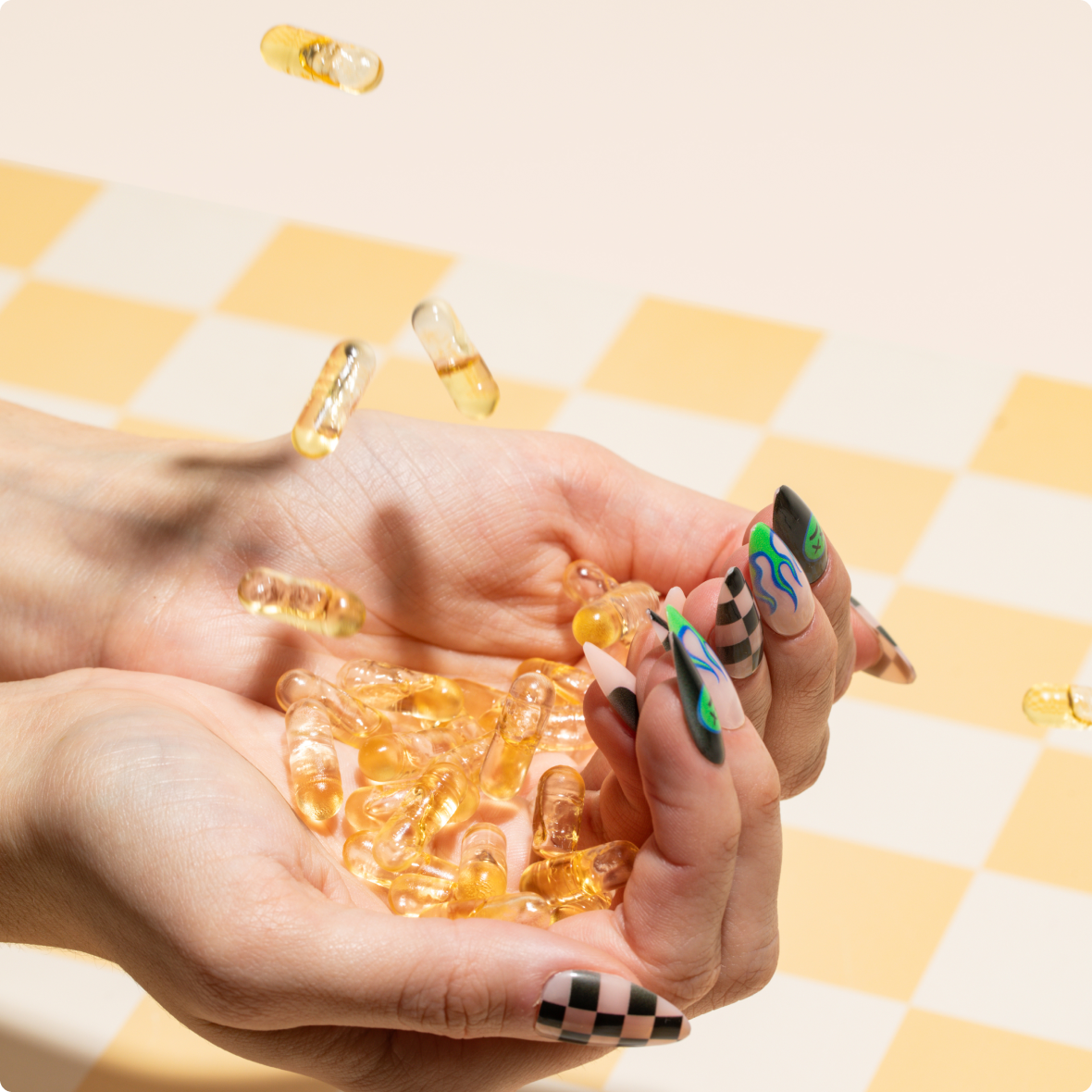 Hands with long decorated nails holding translucent golden gel capsules over a yellow-and-white checkered surface