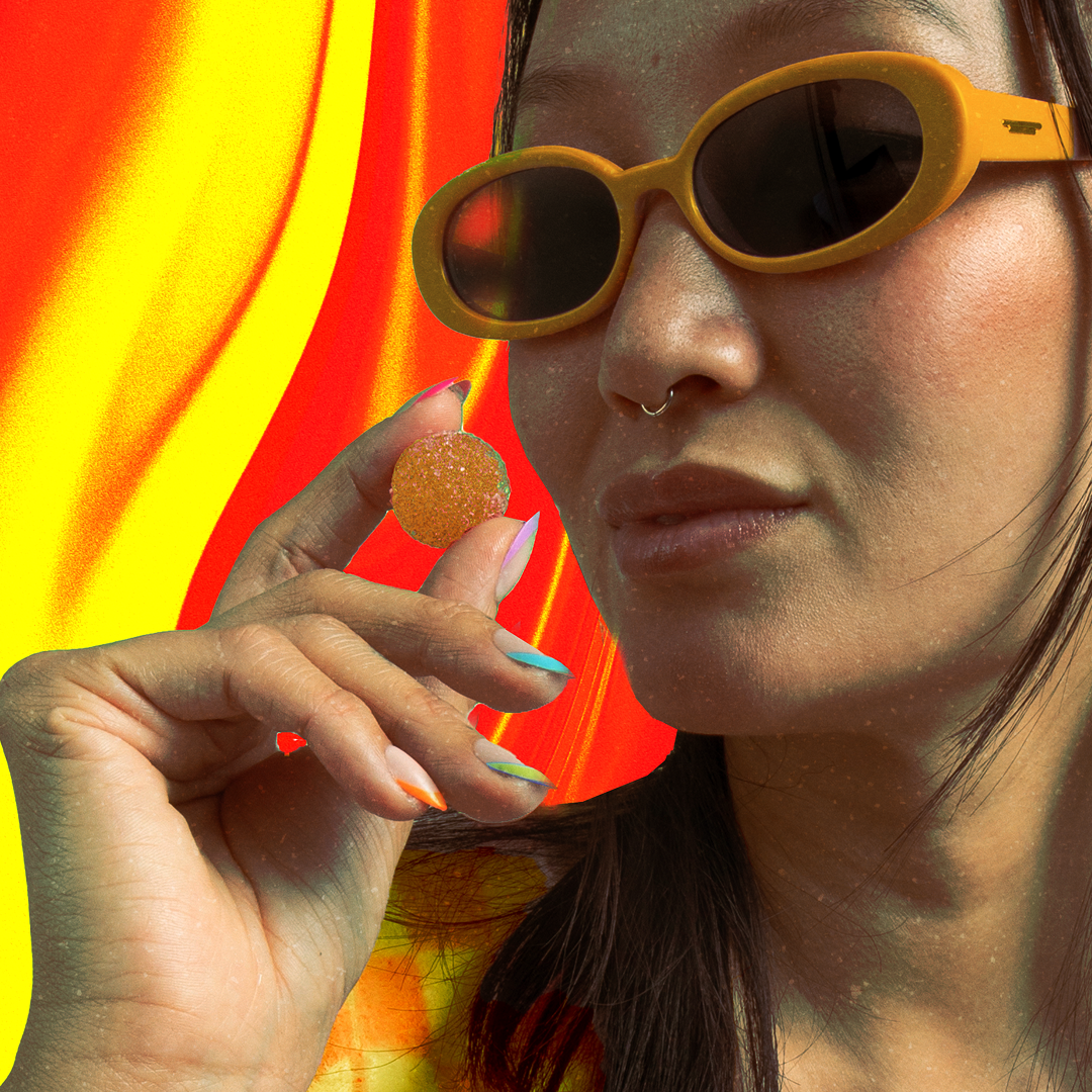 Woman with yellow sunglasses and septum ring holding a sugar-coated gummy between colorful manicured fingers against red-yellow wavy background.
