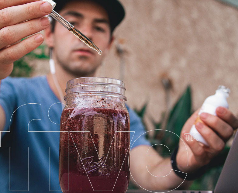 Man adding drops from a dropper into a Ball mason jar of purple smoothie; translucent 'five' overlay with 'TM'.